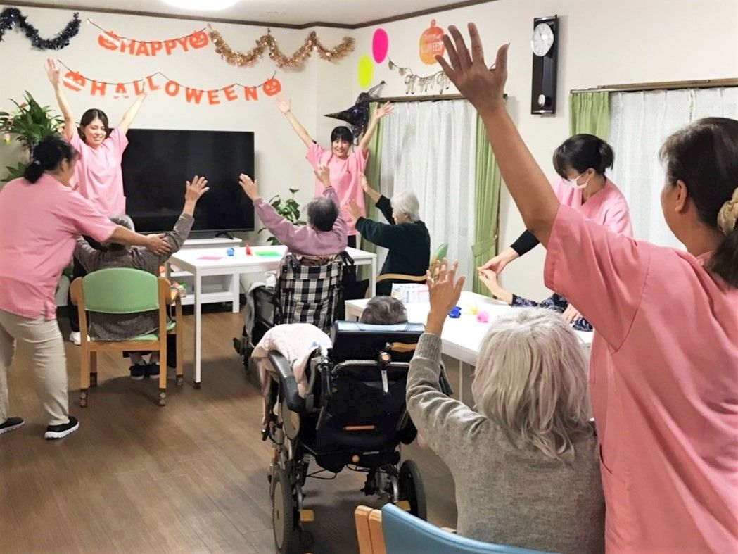 施設のリビングで体操している高齢者の様子。スタッフはピンクの制服を着ている