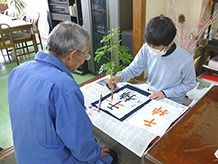 書道に取り組む施設内の利用者とそれを採点しているスタッフの様子