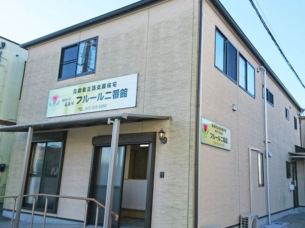 ブラウン系の外壁の2階建ての建物は玄関には手すり付きのスロープがあり、外壁には住宅名が書かれたプレートが貼られている。
