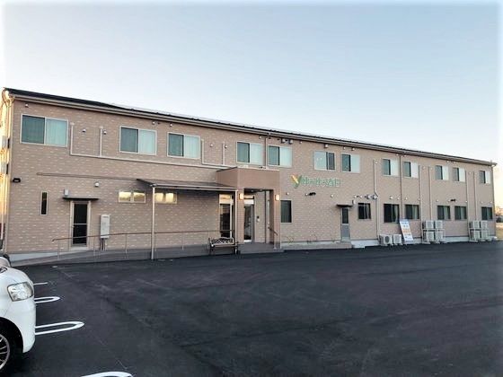 横に長い２階建ての建物の外観写真。手前に駐車場、建物の中ほどにエントランスがある様子