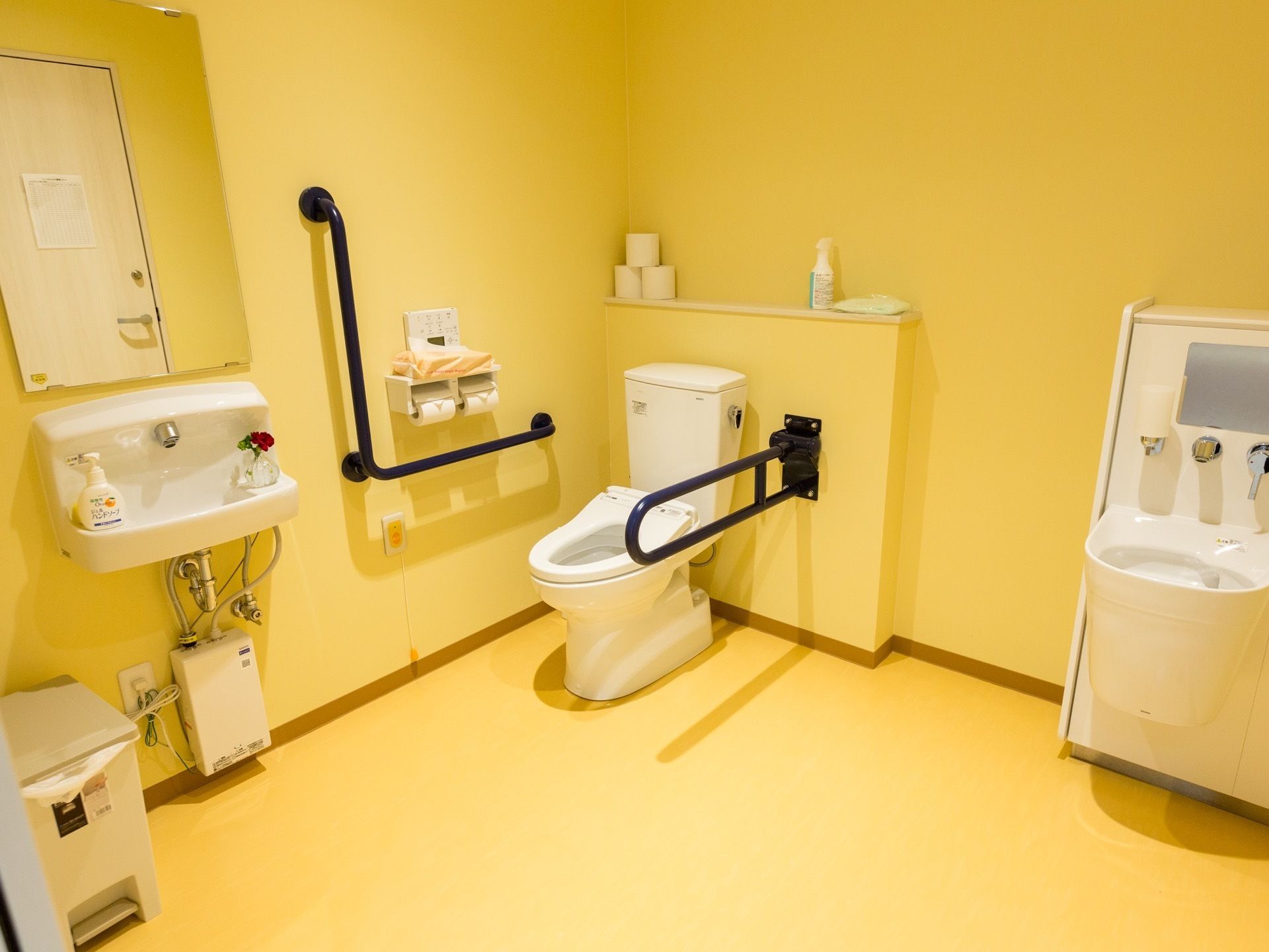 開放的なトイレである。壁と床はイエローで統一されている。壁には手すりが取り付けられており、手洗い場が併設されている。