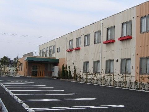 ２階建てで屋根がフラットなボックス型の建物で、前に広い駐車場がある施設の外観