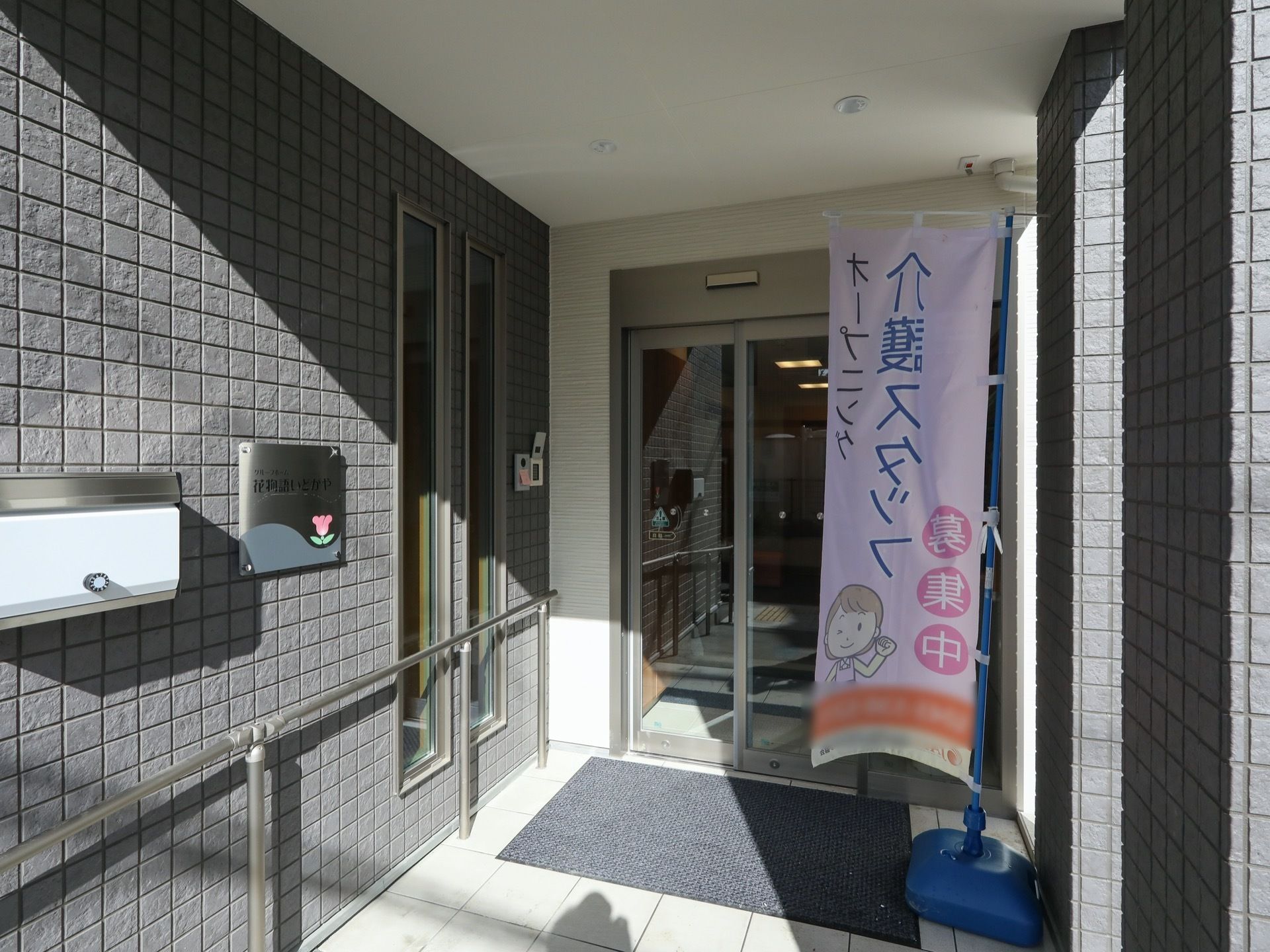 玄関の自動扉の前に足ふきマットが敷かれている。マットの横に、ピンク地の介護スタッフ募集ののぼりが立っている。