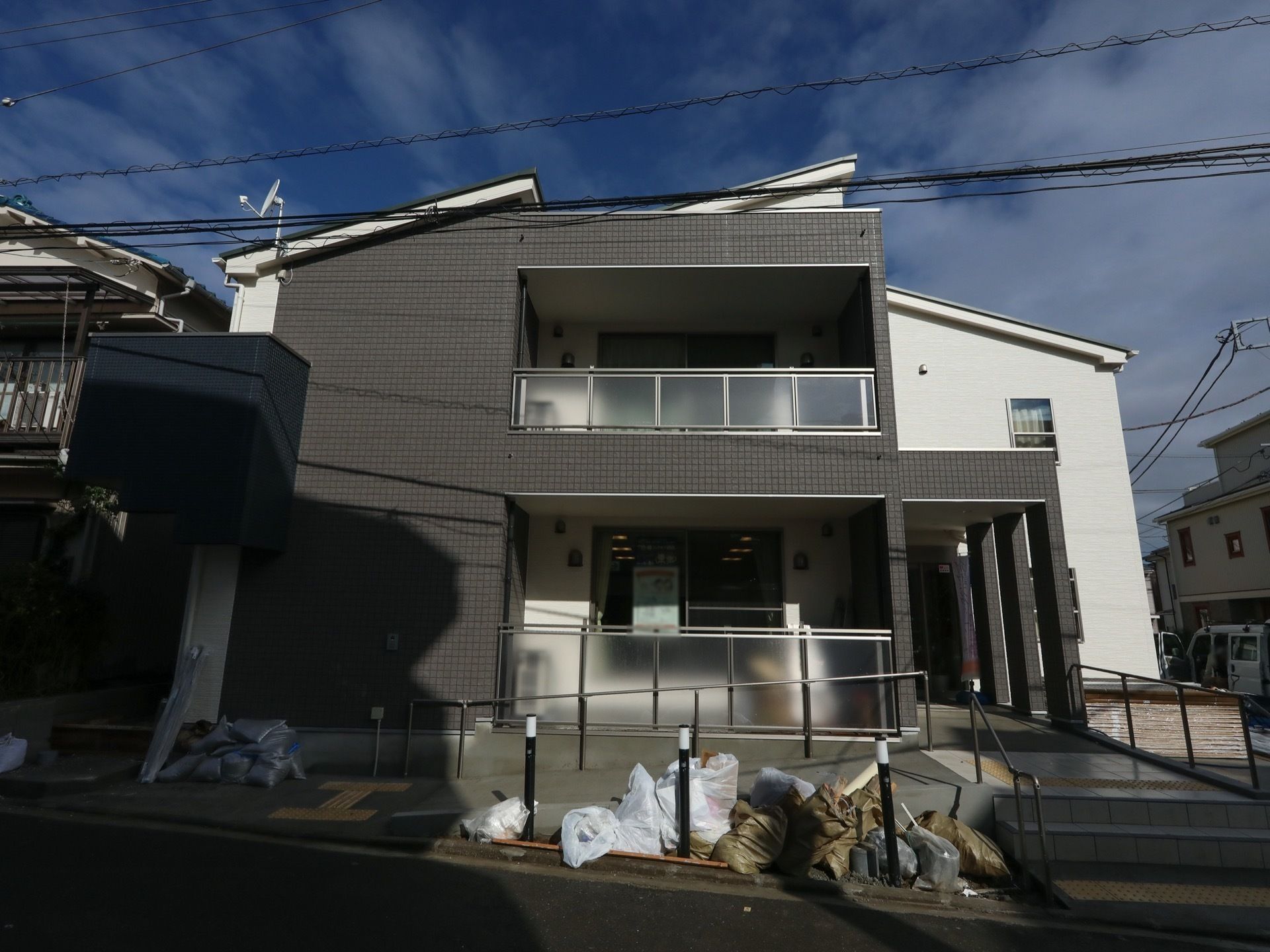 道路沿いに建っている２階建ての施設である。玄関前の道路際にポール状の電灯があり、その周辺にごみ袋が置かれている。