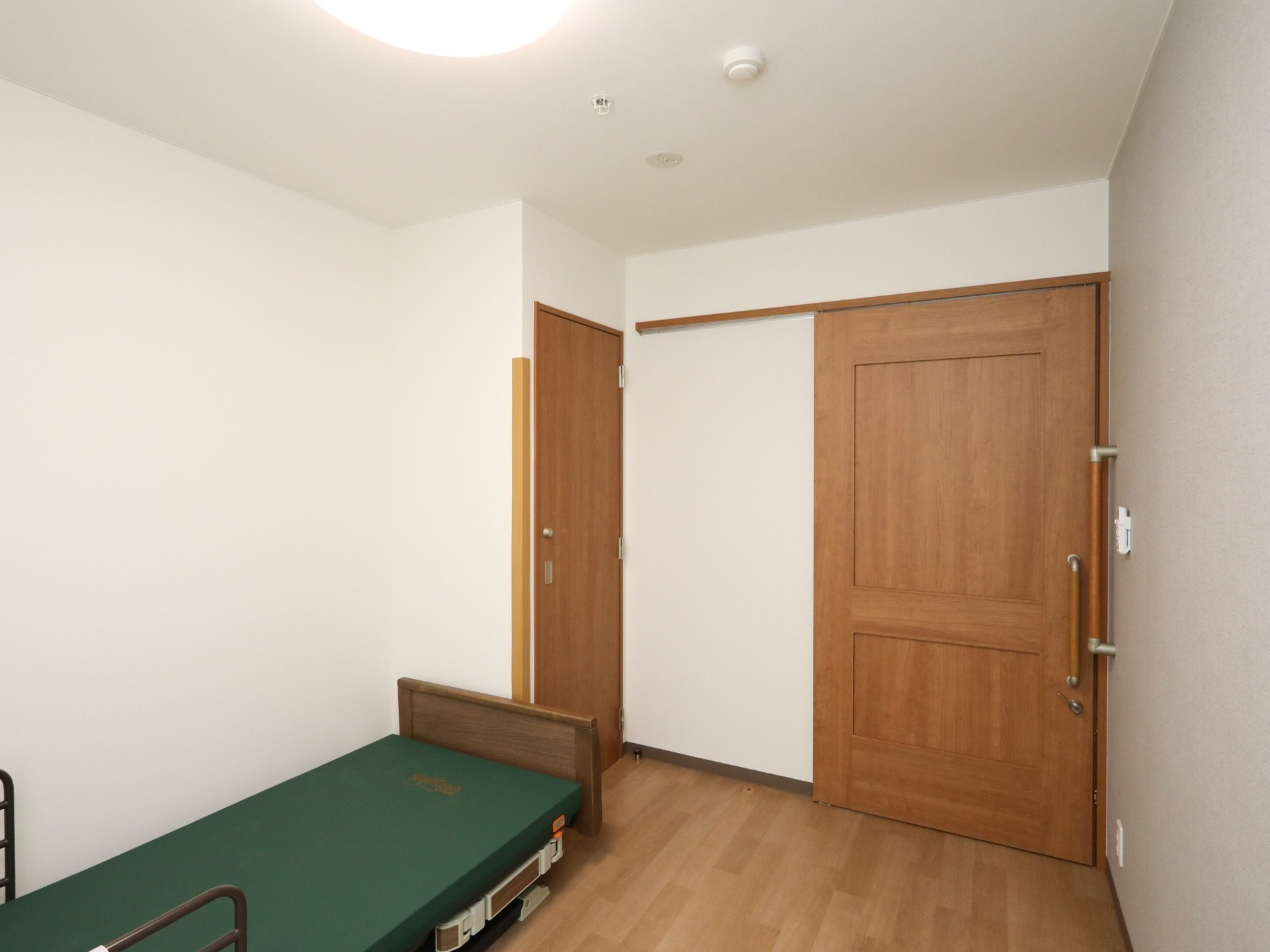 居室入口扉のすぐ横の壁に縦方向の大きな手すりが取り付けられている。天井に煙探知器とスプリンクラーが設置されている。
