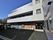 施設の建物の前は駐車場となっている。数台の車が停められている。白の外壁の建物とダークグレーの外壁の建物の２棟が繋がっている。
