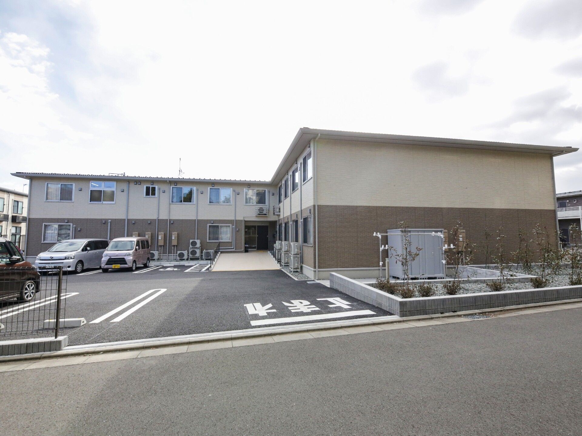 駐車場越しに撮ったブラウンとホワイトのツートーンのＬ字型の建物