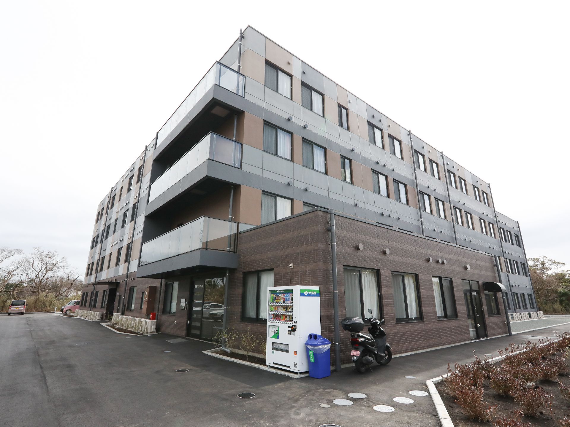 窓がたくさんついた4階建ての建物。周辺には畑や緑があり、青空が広がっている。