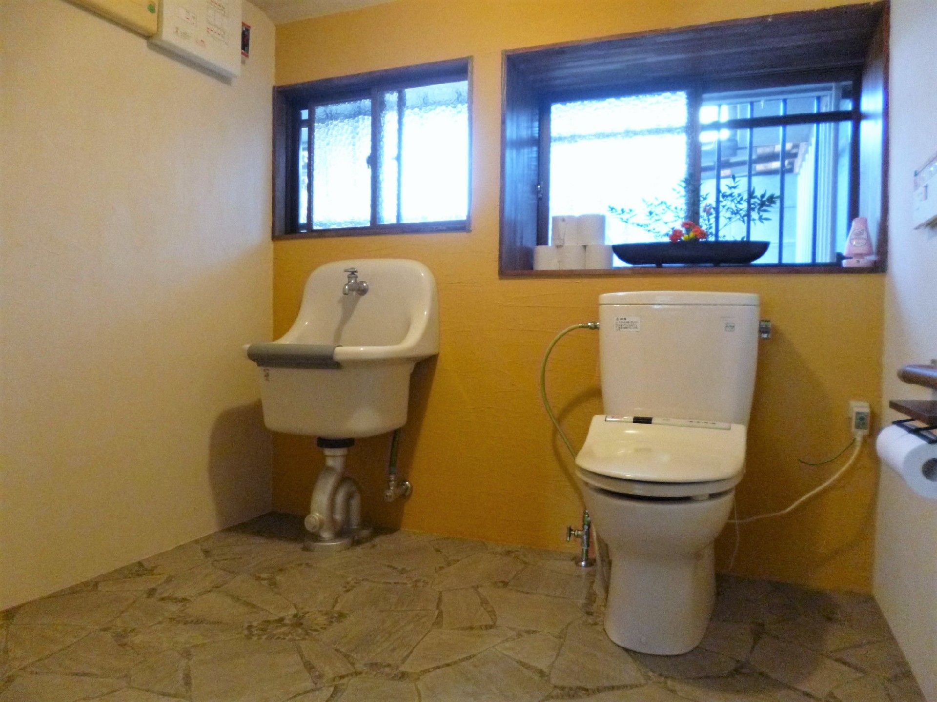 広いトイレの右側に様式の便座が置かれ、左側に洗い場があり、大きな窓のそばにはトイレットペーパーが積まれている。
