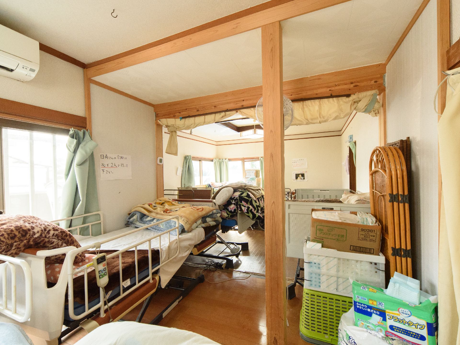 壁に介助の際の注意書きがあり、複数の介護用ベッドと予備の毛布やおむつが置かれた部屋