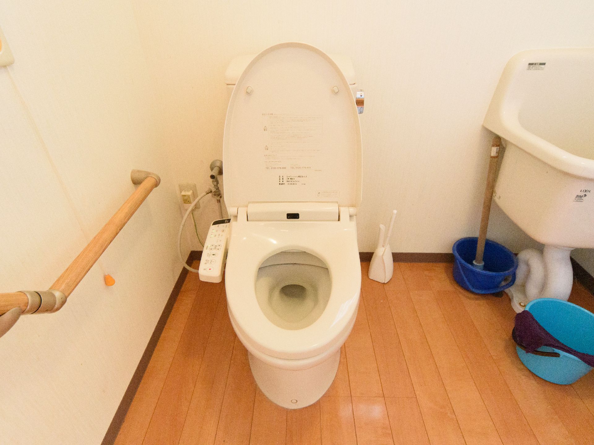 手すりが設置され、深い洗面台と掃除用具が備えられたトイレの個室
