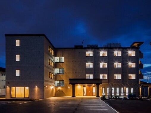 4階建てのエル字型の建物の玄関には車寄せが付いている。周囲が暗くなり、玄関がライトアップされ、窓から明かりが漏れている。