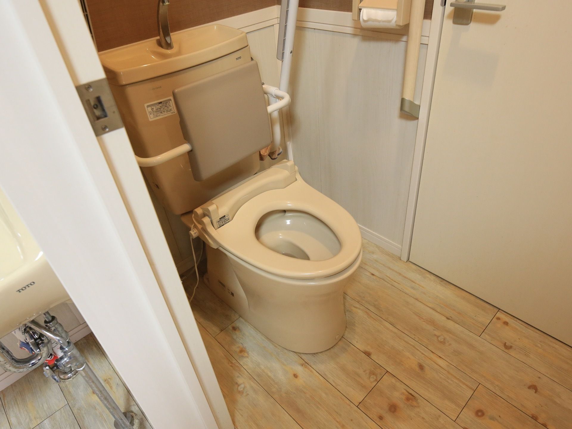 淡いライトブラウンの木目調の床が温かみを与えるトイレである。便座にはクッション性のある背もたれが完備されている。