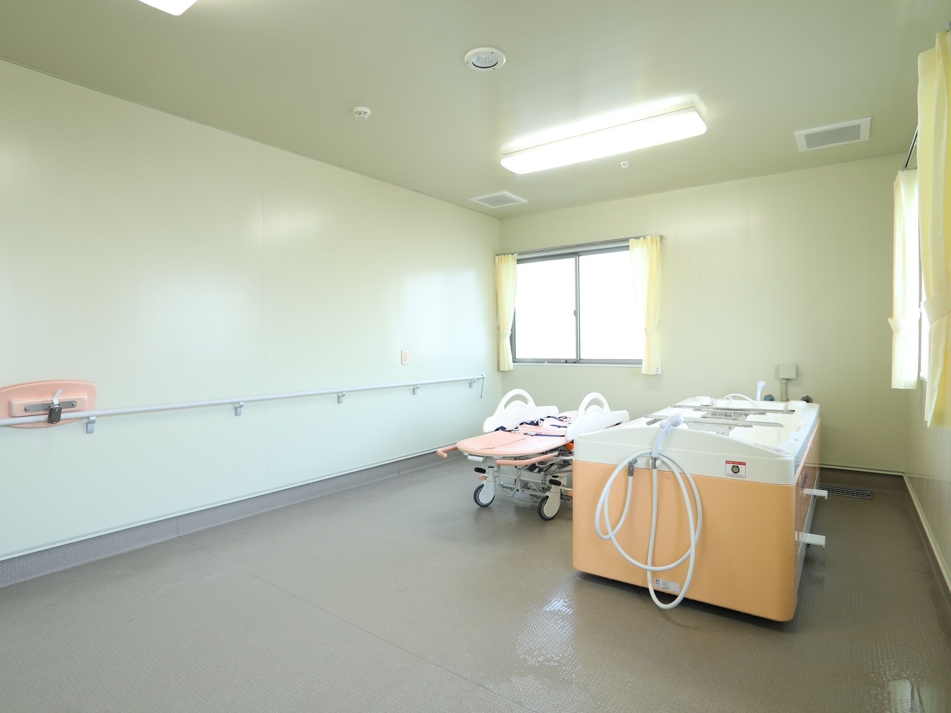 大きな窓が設置された、機械浴室がある。床はグレー。壁は白を基調としている。リフターや浴槽が置かれている。