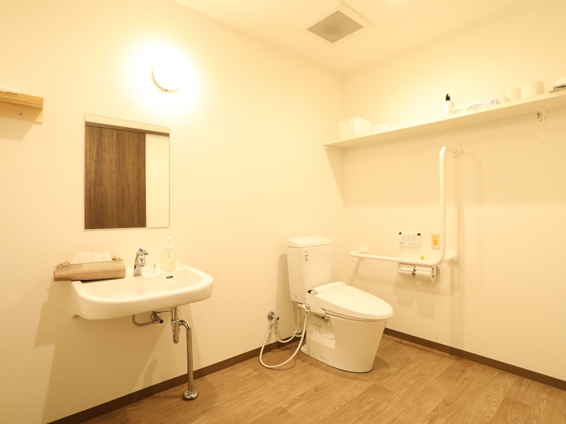 上側に収納スペースを完備した、広い洋式トイレである。壁側に手すりががある。すぐ近くに洗面台もある。