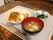 白いトレイの上に、スパゲッティと揚げ物の皿、澄まし汁、青菜の和え物の小鉢がのっている。手前には箸とフォークが並んでいる。