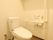 白い壁と木目調の床の個室のトイレがある。天井には棚があり、壁には手すりが設置されている。温水洗浄便座が付いている。
