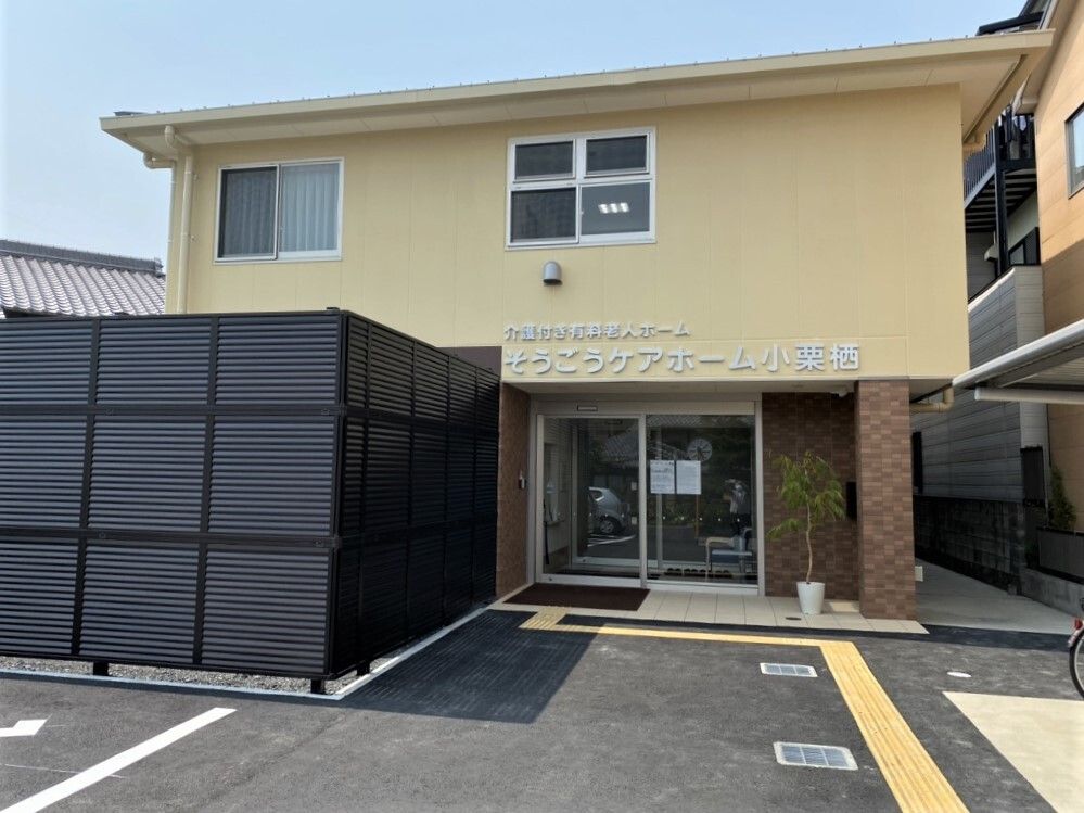 ベージュを基調とした2階建ての建物の外壁にはホーム名が書かれ、黒い柵で囲まれたスペースもある。