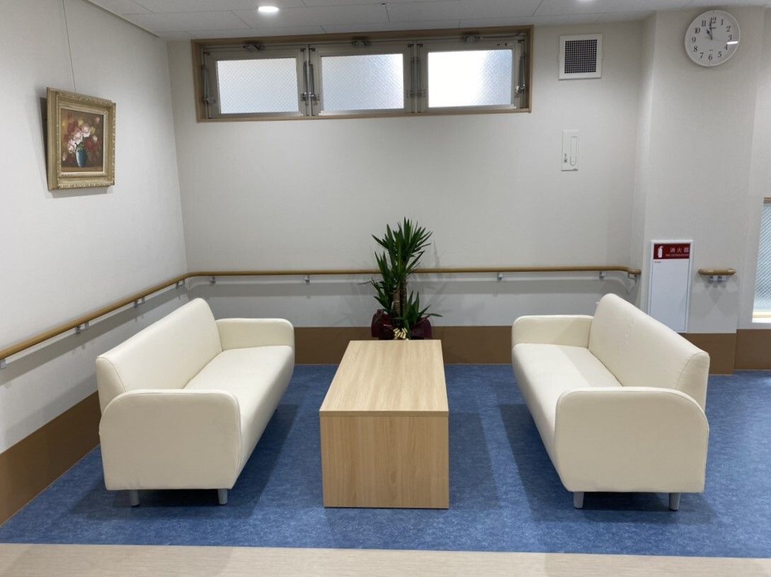 白いソファや木製のテーブルが置かれた共有スペース。観葉植物や絵画も飾られ、壁には手すりがある。