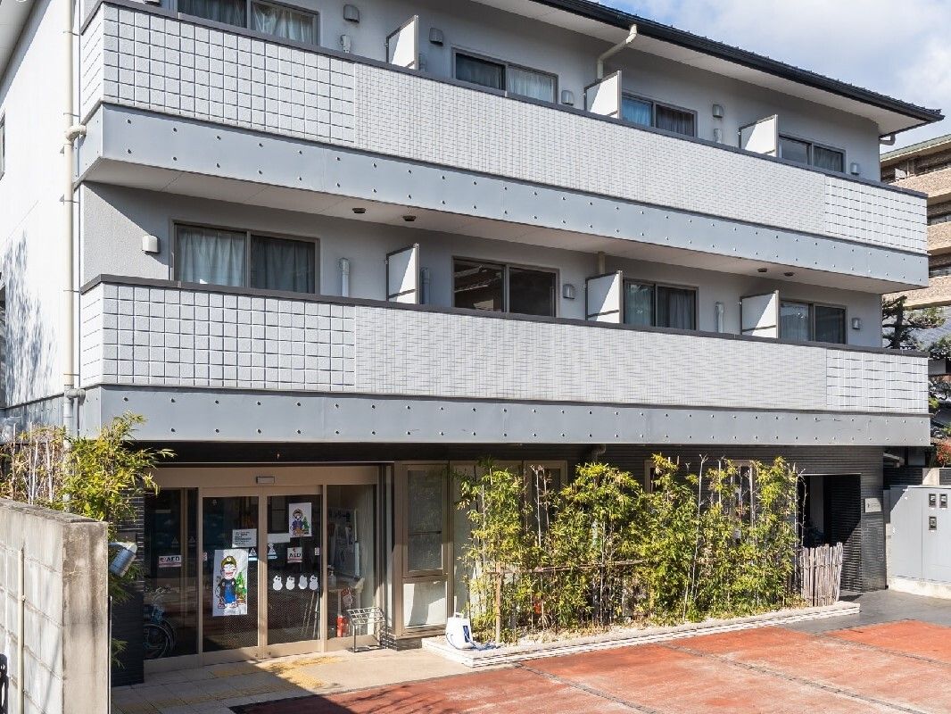 3階建てのグレー系の建物。建物のある敷地は広く、ベランダも付き、周囲には緑もたくさん植えられている。