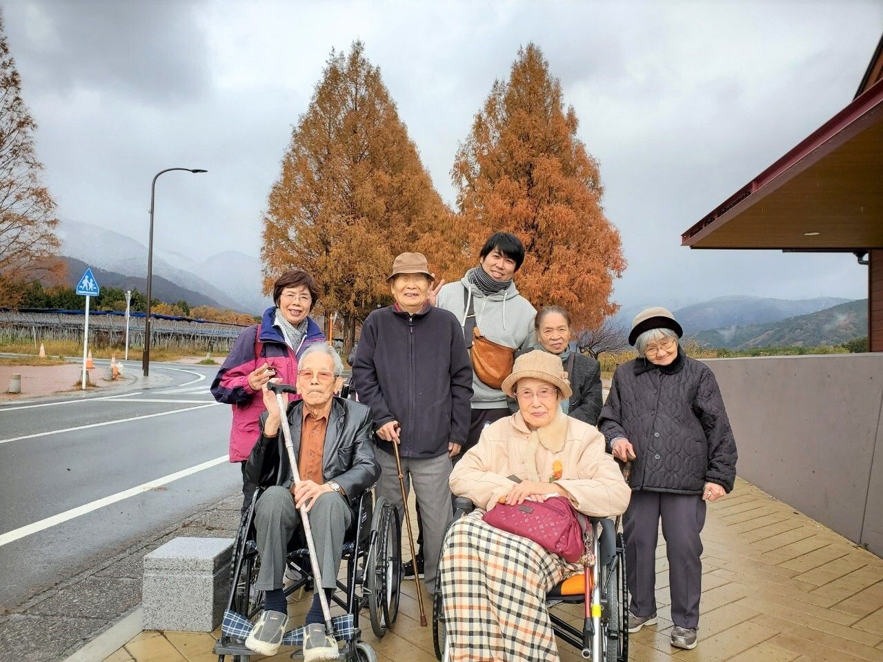 山に囲まれた景色の中の歩道で、記念撮影に応じる入居者の方々とスタッフ