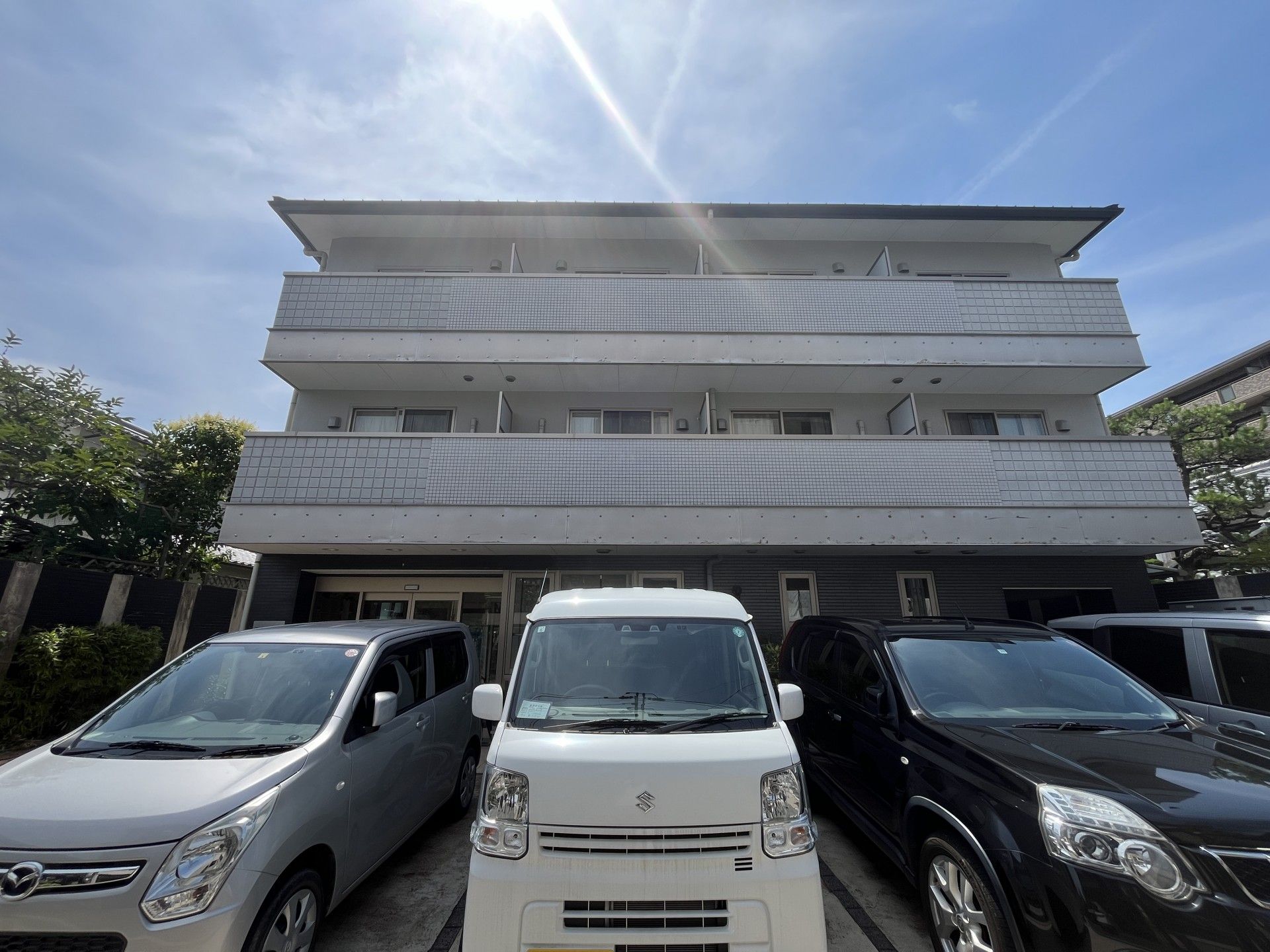 グレー系の３階建ての建物。周辺には木が植えられ、玄関には自動ドアが設置され、傘立てもある。