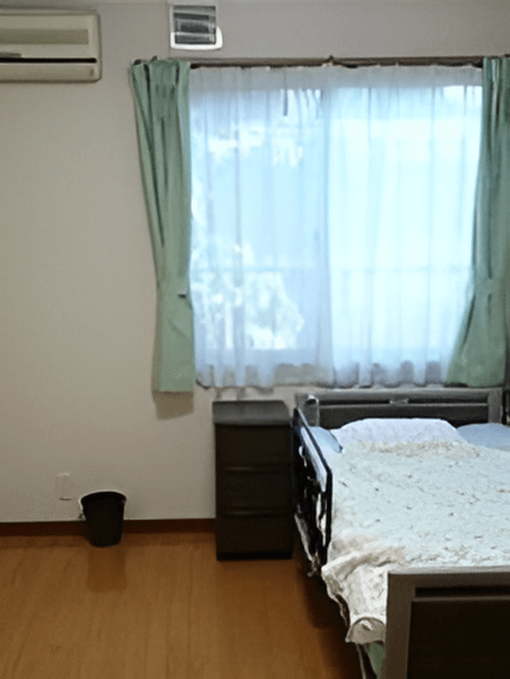 寝具付きのベッドやゴミ箱、エアコンが設置されたフローリングの部屋にはレースカーテンとカーテンが付いた窓がある。