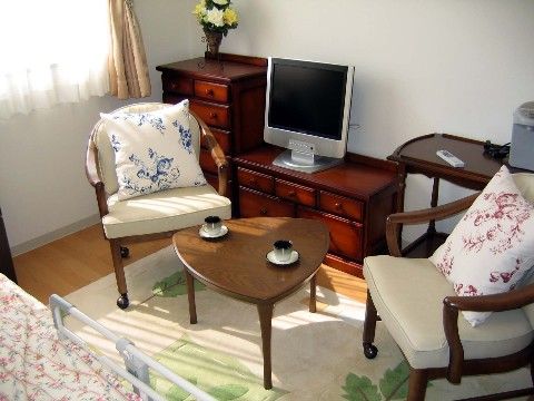居室の床はフローリングで、ベッド、テーブル、椅子、タンス、テレビが設置されている。壁の色は白色基調で、窓が設けられている。