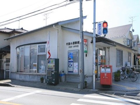 地域の郵便局の建物である。壁の色は白色系で、局名が表示されている。玄関の前には郵便ポストが置かれている。