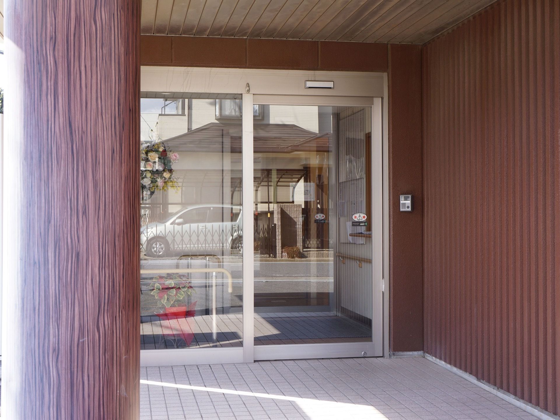 玄関のドアは幅広の自動ドアになっており、すぐ脇にインターホンが設置されている。ドアガラスの奥には花や鉢植えが置かれている。