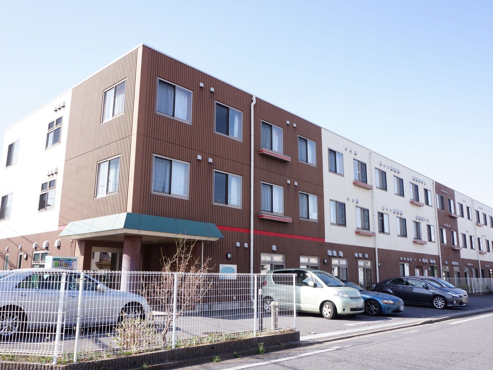 外壁が茶色と白の３階建てで、建物の前に車が多数駐車している施設の外観