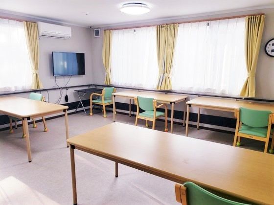 長机と椅子が並んでいる施設内のリビングの様子。テレビが壁に掛けられている空間