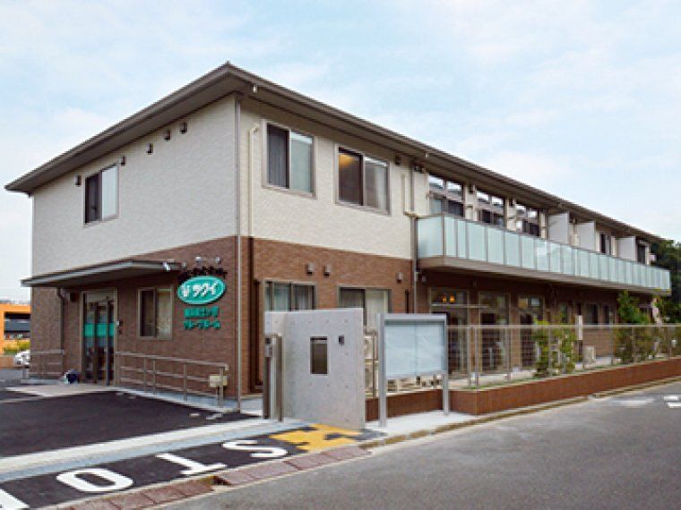 白とブラウンの外壁の２階建ての建物は日当たり良好で敷地内には駐車場があり、敷地の入り口にはSTOPと書かれている。