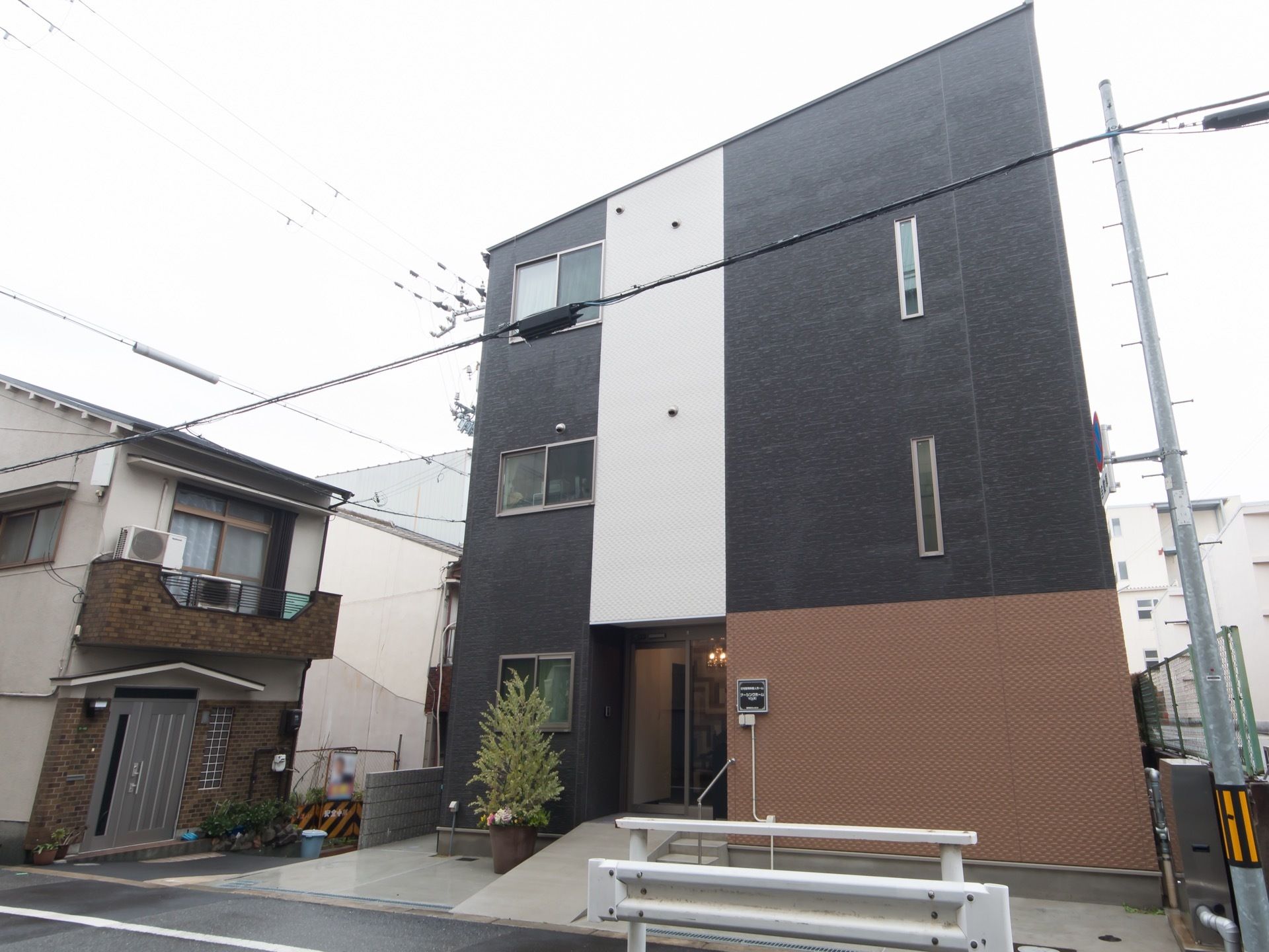 黒と白と茶色の壁が特徴的なシンプルな外観をしている。玄関まではスロープになっており、階段部分には手すりが設置されている。