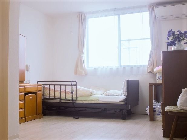 エアコンが付いたフローリングの居室。一人用の椅子や鏡台、寝具付きのベッドがある。