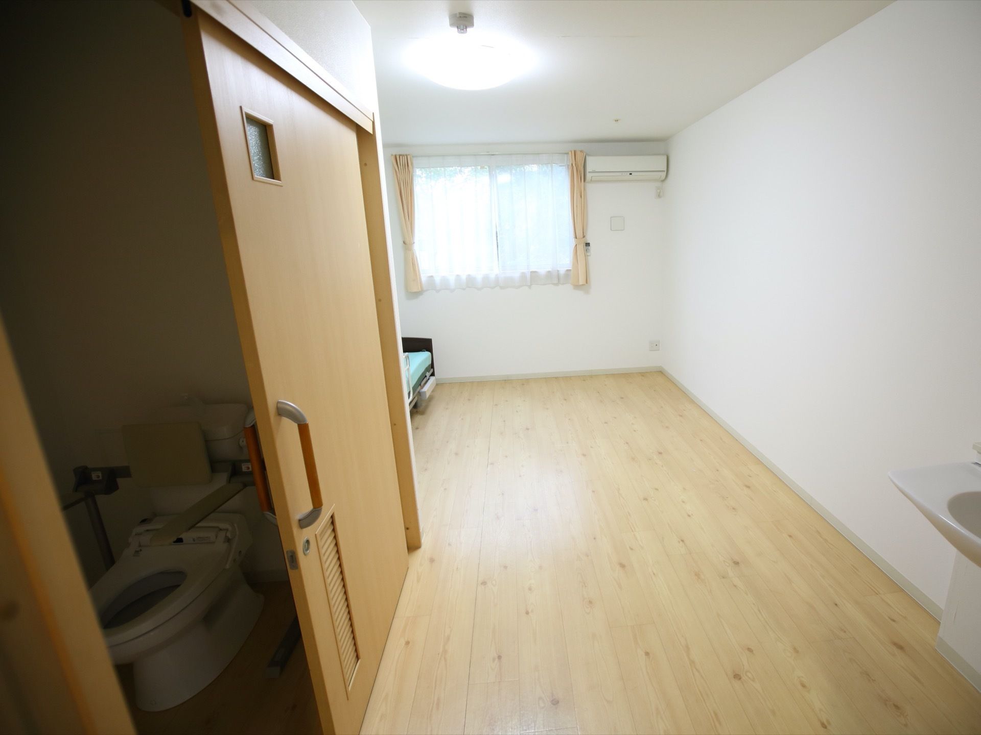 居室の床はフローリングになっており、白を基調とした明るい空間である。左側にはトイレがあり、引き戸式のドアが設置されている。