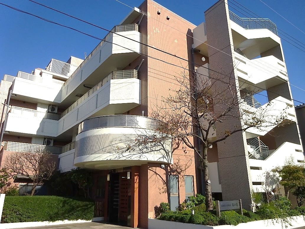 白とブラウンの４階建ての建物の周りには緑も多く、屋外階段やベランダがある。