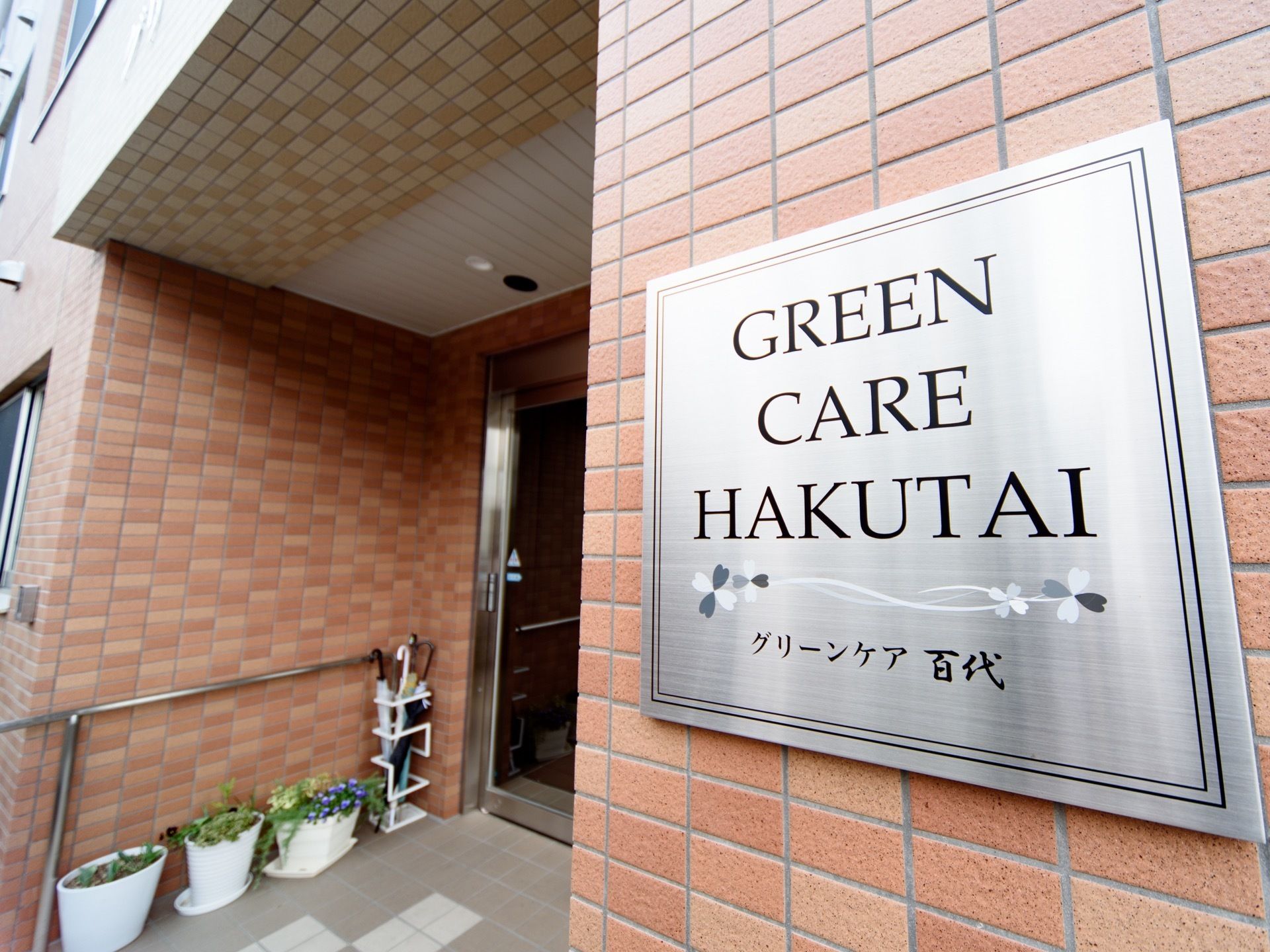 施設はレンガ調の外壁で、シルバーの表札が設置されている。入口には、ガラス張りの自動ドアが取りつけられている。