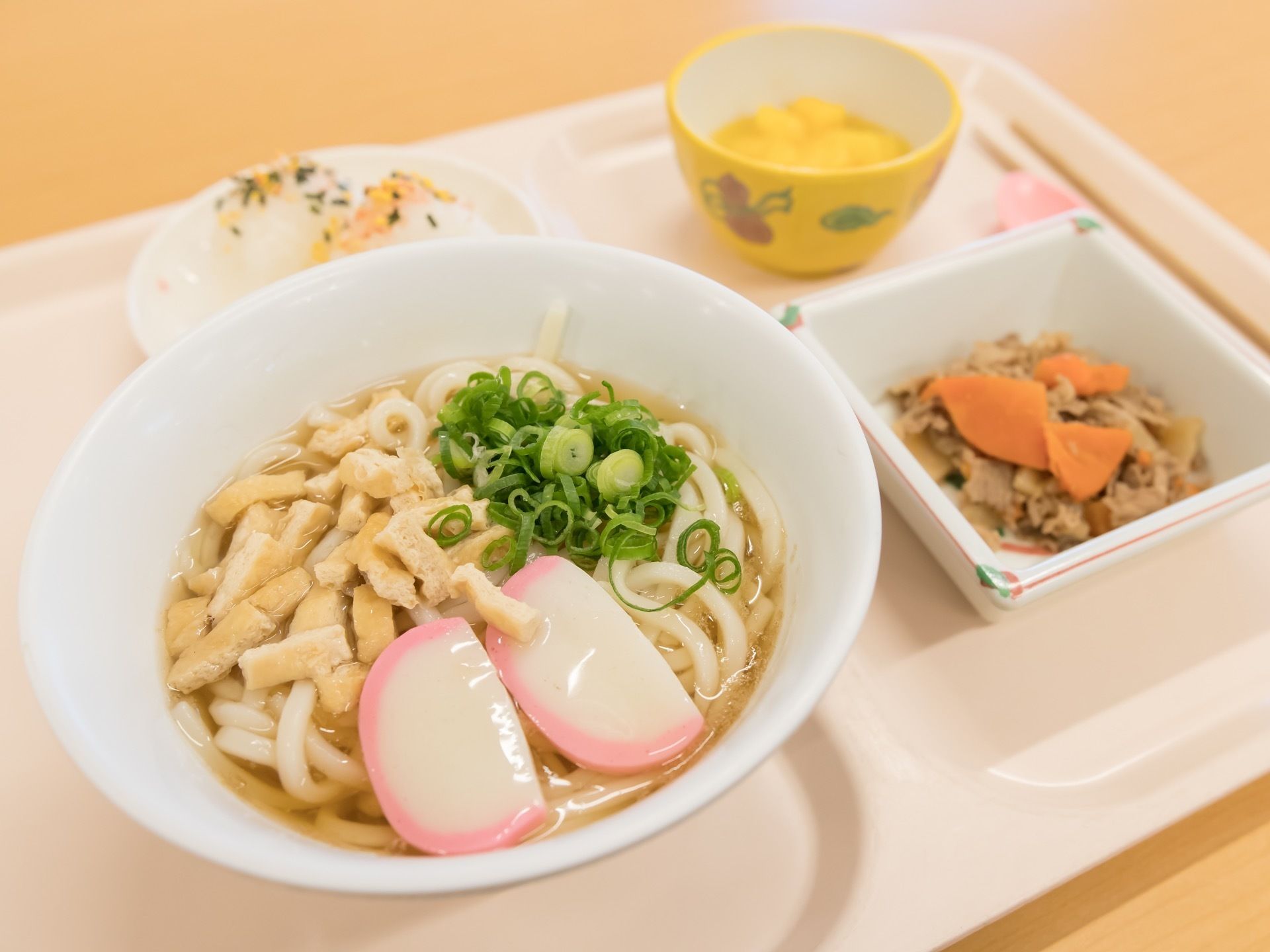 細かく切った油揚げやネギがのったうどんがある。副菜やデザートなどもあり、バランスの良い献立になっている。