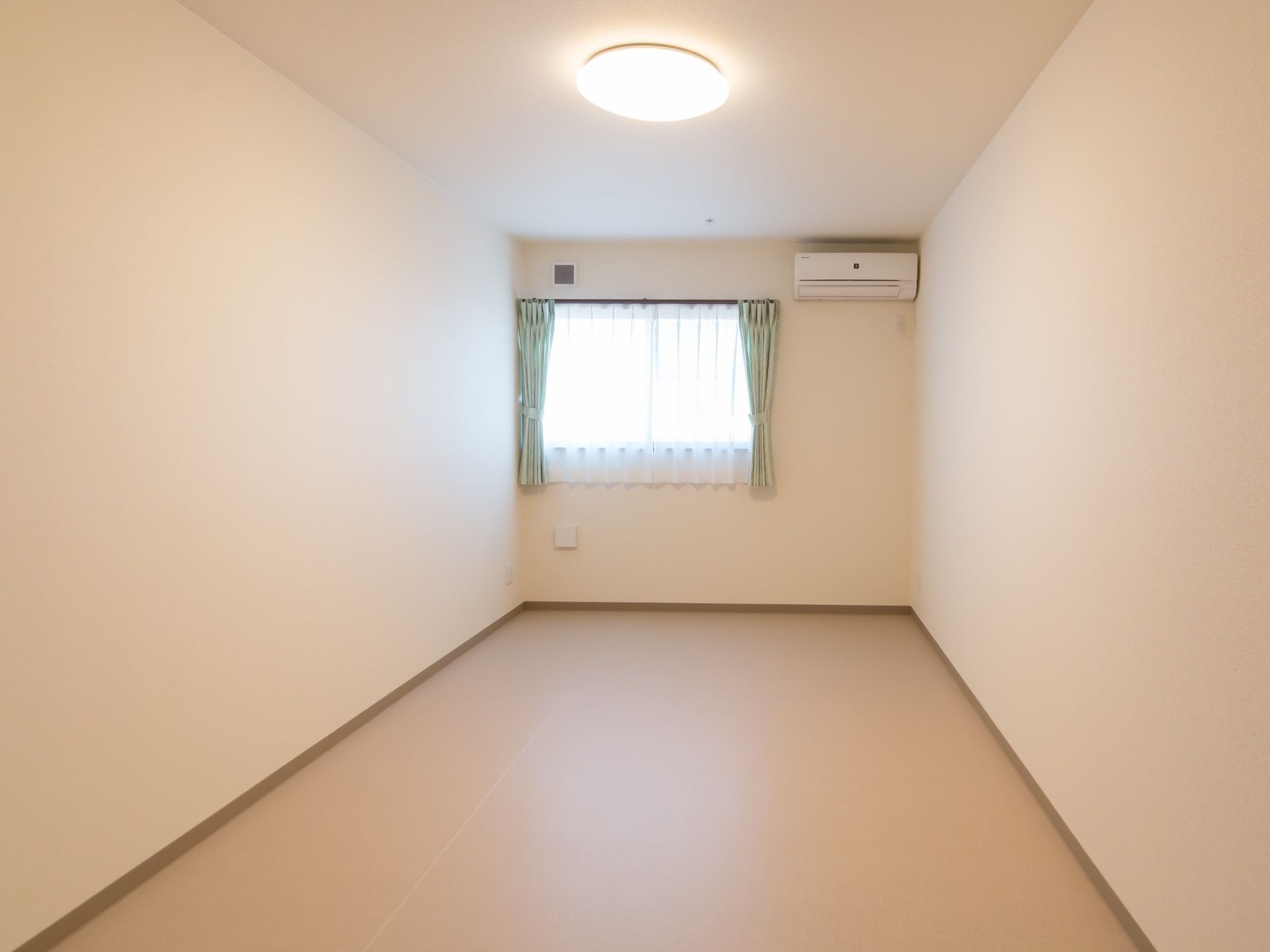 エアコンが完備されているワンルームの居室で、床はフローリングになっている。窓にはレースとカーテンが取り付けられている。