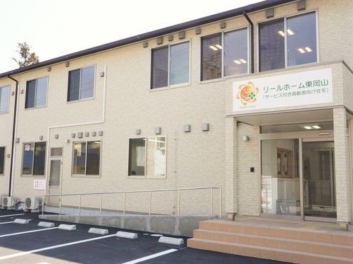 2階建てのブラウンの建物は横に長く窓がたくさんある。目の前には駐車場もある。