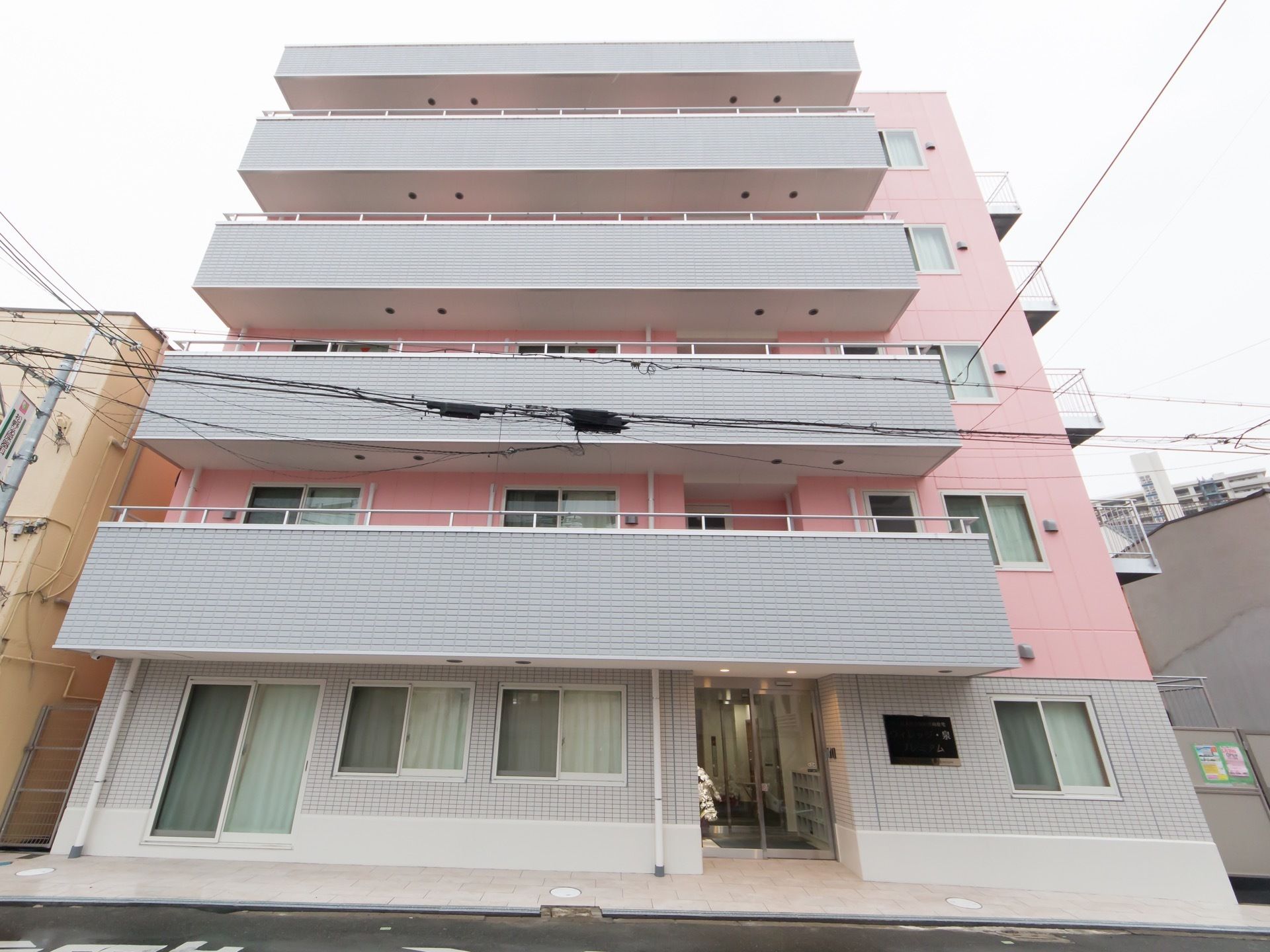 施設の外観は、ピンクとグレーの明るいツートンカラーが印象的になっている。5階建てで居室はベランダに面して造られている。