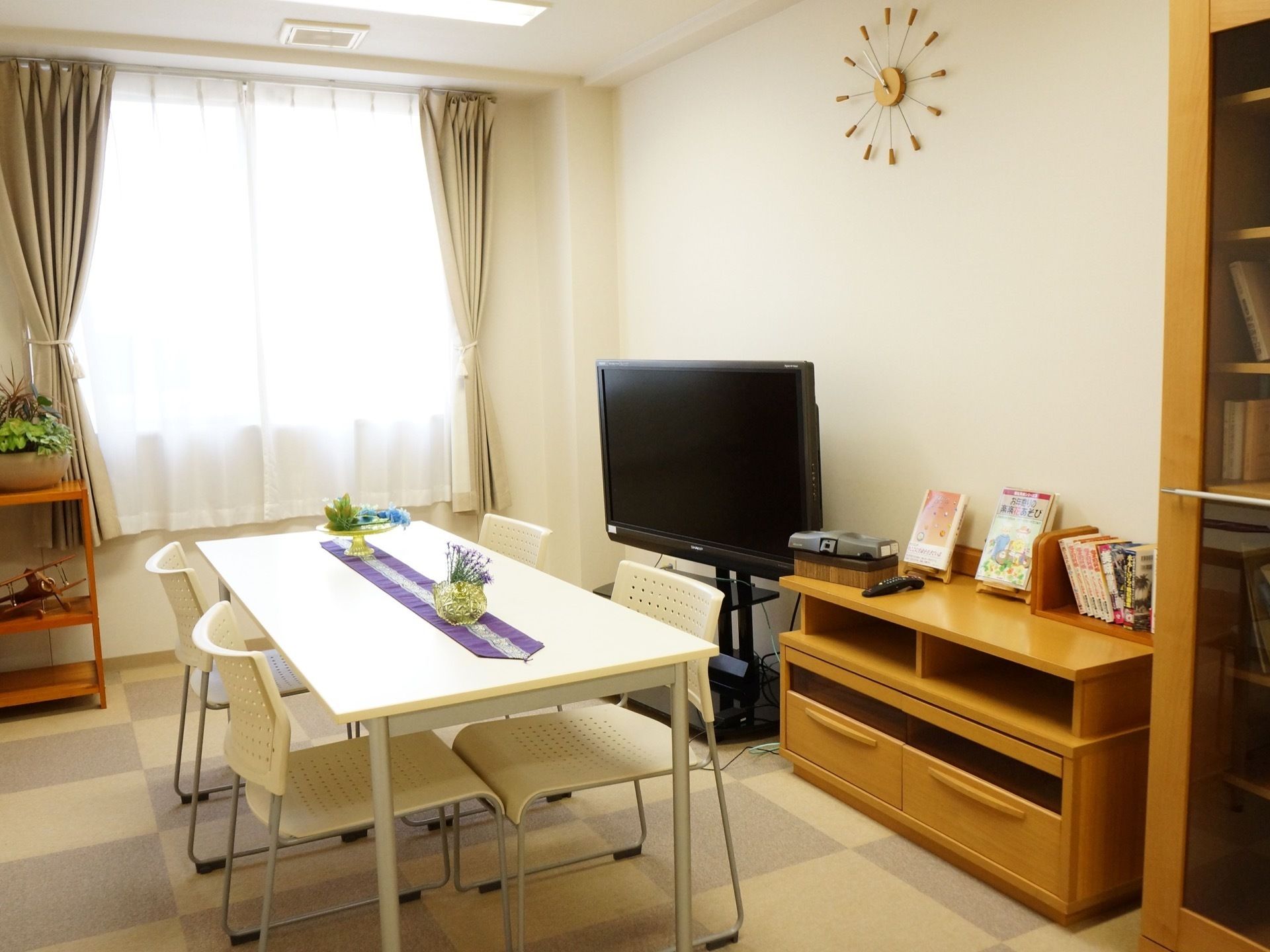 白いテーブルに４脚の椅子が向い合せで置かれている。壁側にはテレビ、本などが置かれている。格子状の絨毯を敷いている。