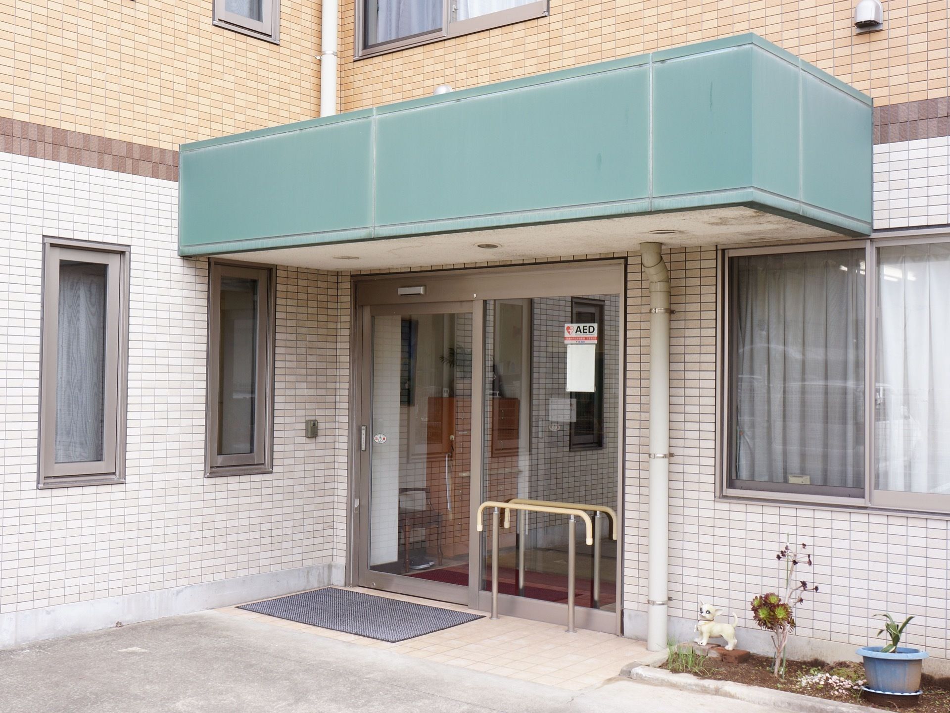 玄関前は広く、バリアフリーになっている。扉の横に手すりがある。AEDのシールが扉に貼られている。近くには花壇がある。