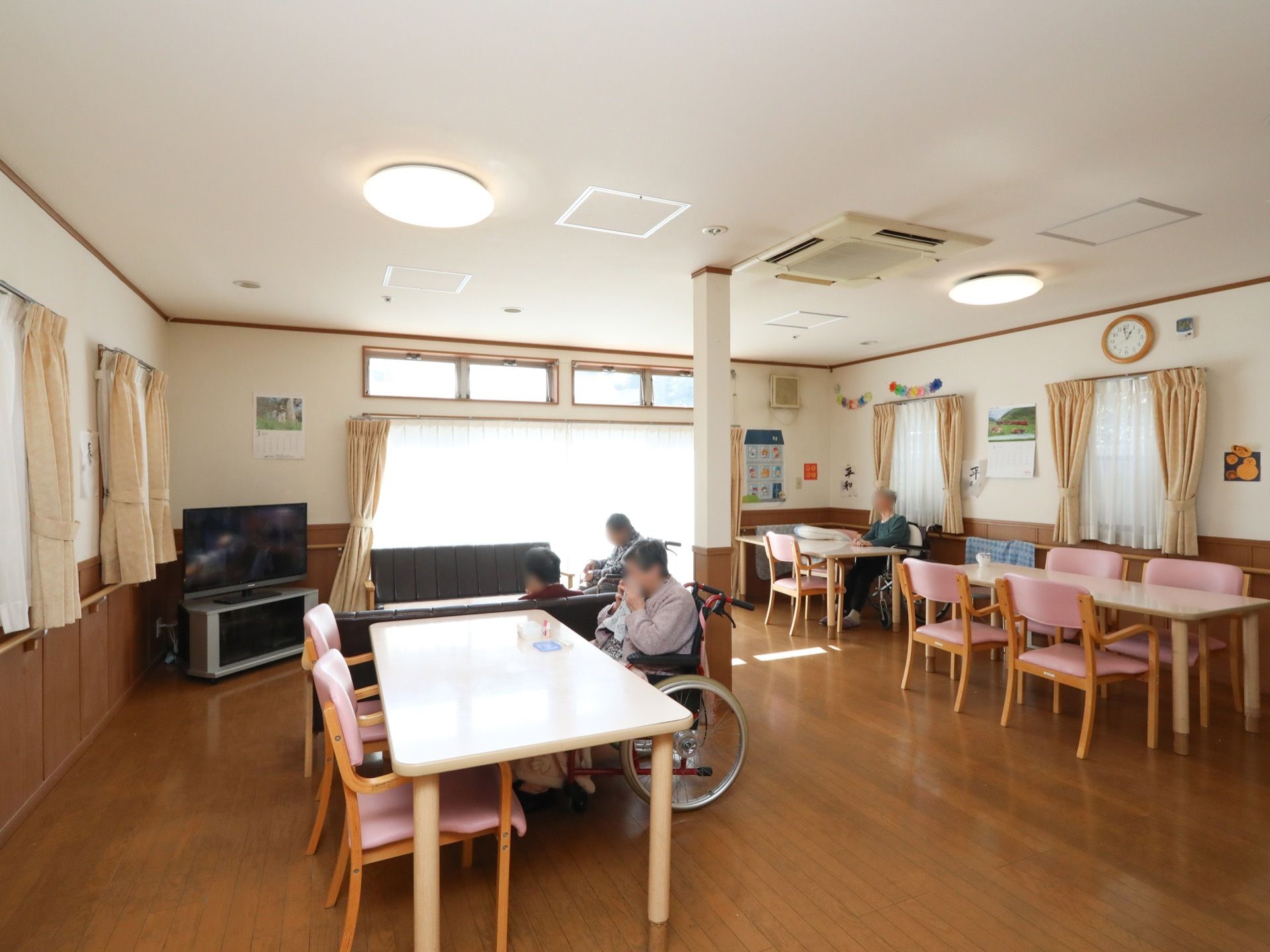 大きな窓のそばにテレビが設置され、テーブルに淡いピンクのシートの椅子が配置された共有スペースである。