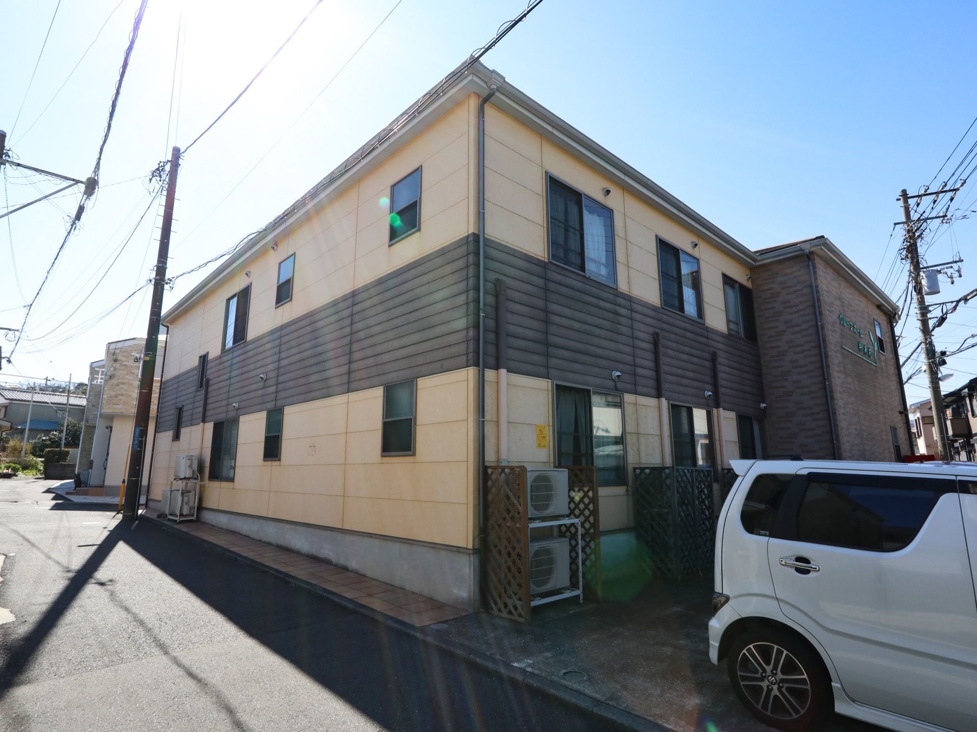 晴天の日に2階建ての施設建物を駐車スペースを右側に対角線状に見た様子である。室外機に日差しよけが設置されている。