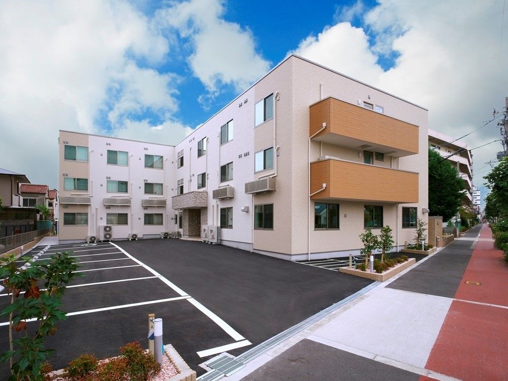 ３階建てでエル字型のベージュ系の建物。周辺には緑も多く、住宅も点在している。