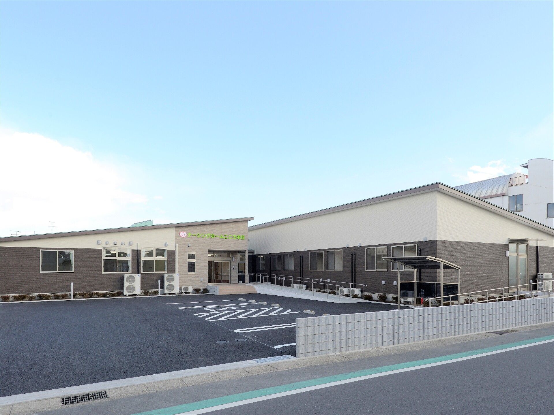 広い敷地内に駐車場と２階建ての建物が建っている様子。天気が良い日の外観