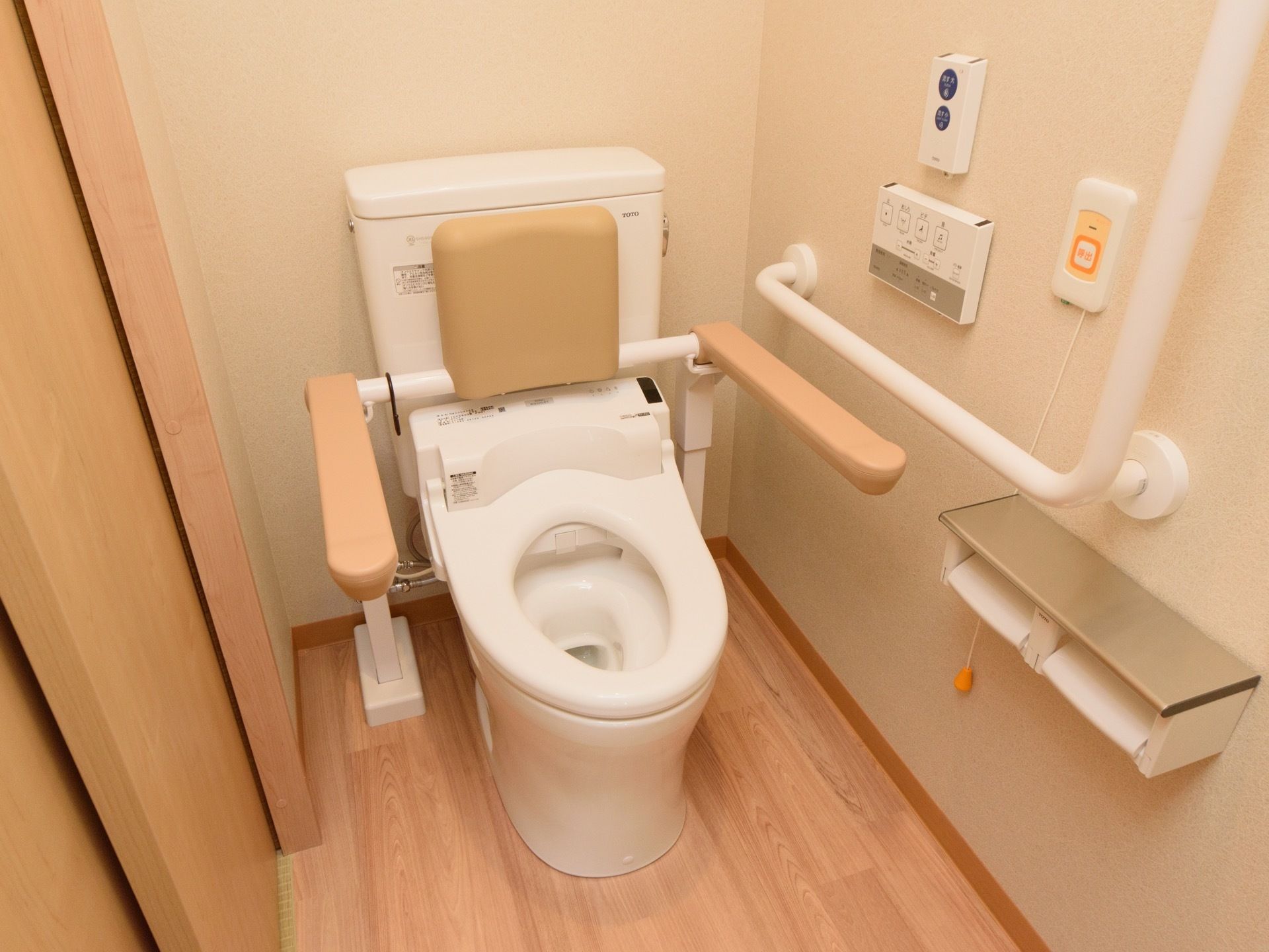 通常サイズのトイレに、背もたれ付きのひじ掛けやＬ字手すりが用意されている。壁には操作スイッチのほかナースコールが設置されている。
