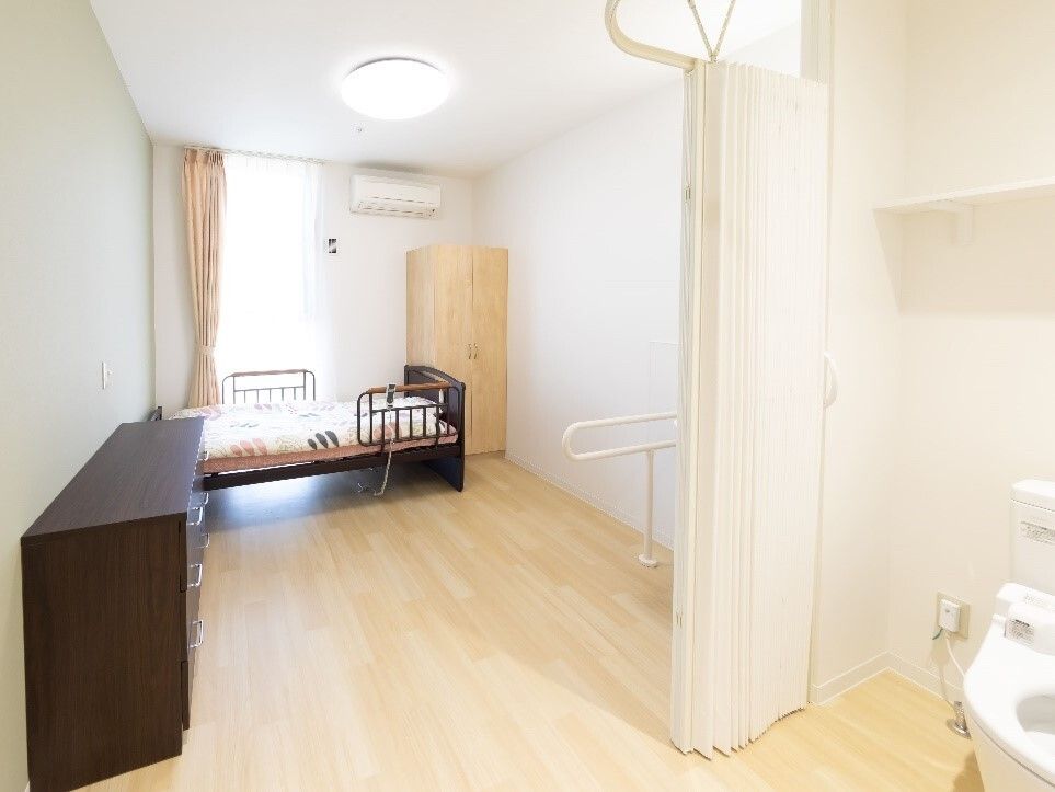 カーテンで開け閉めをするタイプのトイレが手前にあり、奥の窓のそばには介護用ベッドやクローゼットが置かれ、トイレのそばにタンスが設置されている。